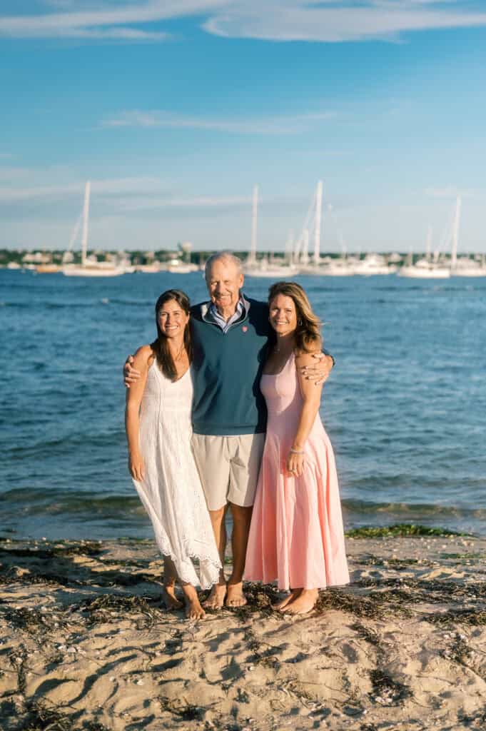 Three people stand barefoot on a sandy beach at Brant Point, posing for photographer Kaity Farrell with boats and water in the background on a sunny day.