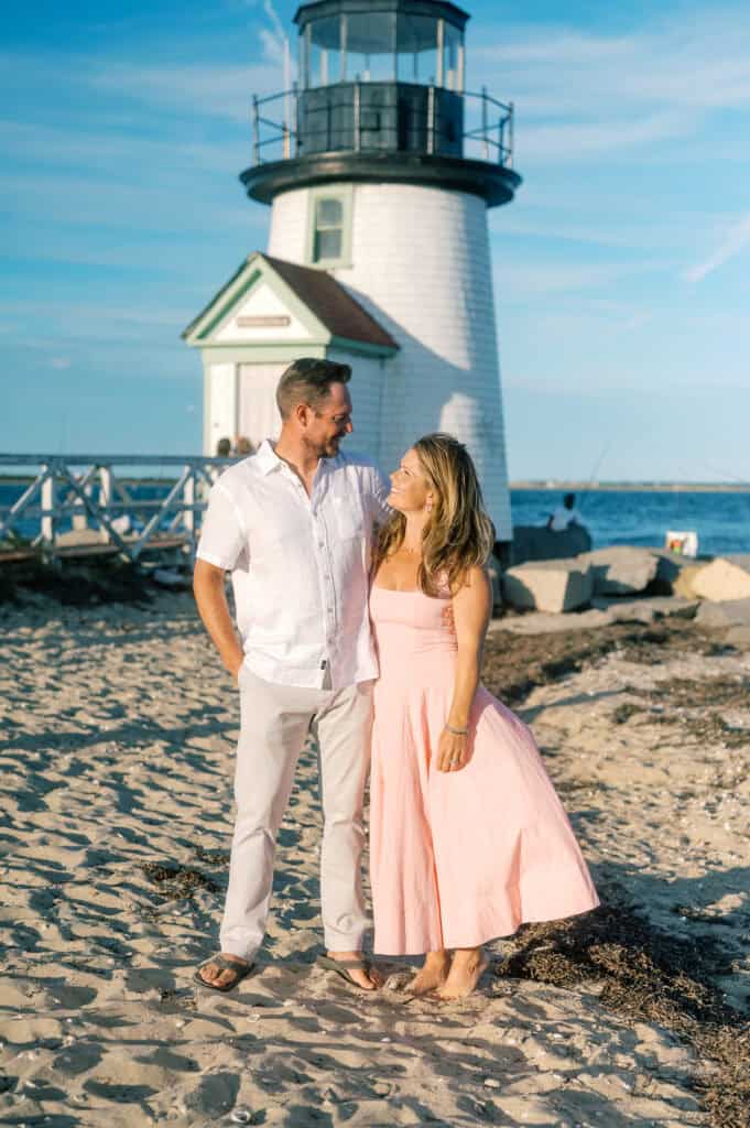 A man and woman stand together on a sandy beach in front of a lighthouse, looking at each other and smiling—captured by photographer Kaity Farrell during their Nantucket vacation family photos.