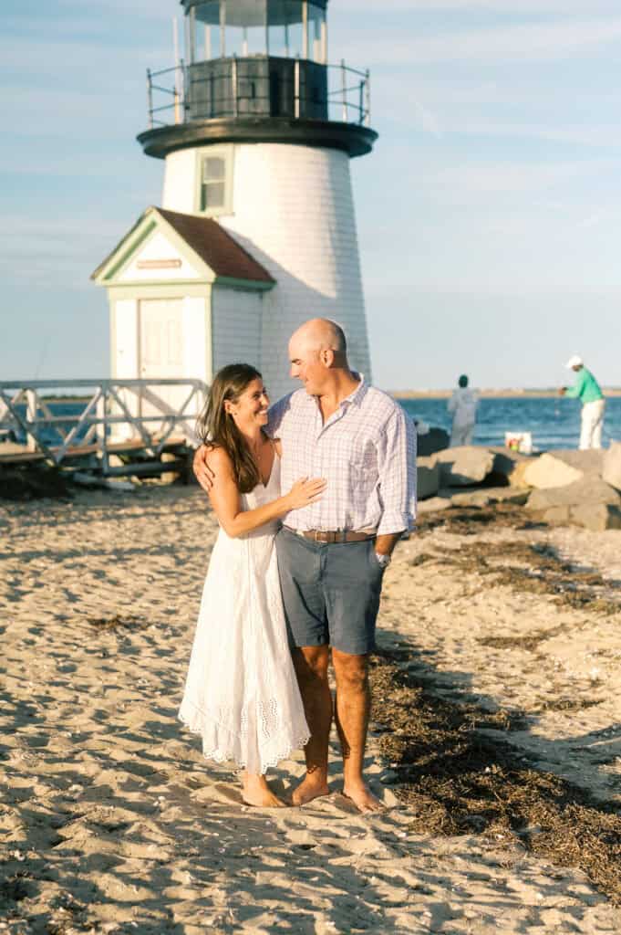 A couple stands on a sandy beach near a lighthouse, smiling at each other, while a person fishes on the rocks in the background—captured by Nantucket family photographer Kaity Farrell.