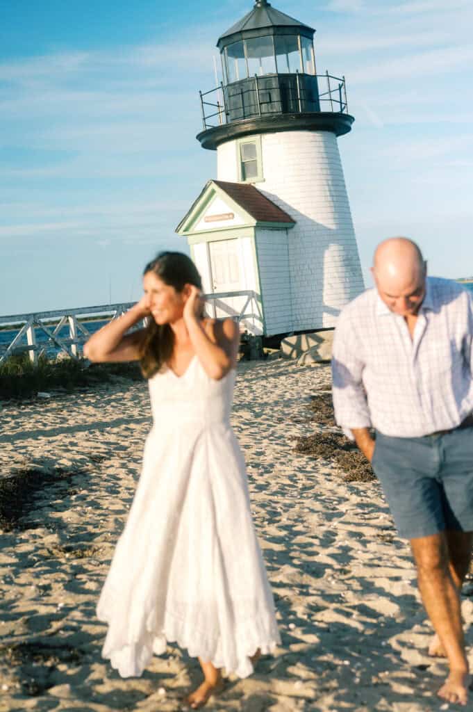 A woman in a white dress and a man in casual clothes walk on a sandy beach near Brant Point Lighthouse under a clear blue sky, capturing special moments perfect for photographer Kaity Farrell’s family photos.