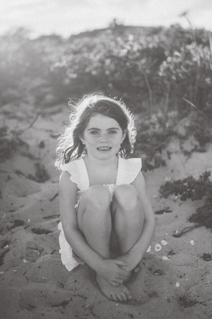 Young girl in a white dress sits on sandy ground with her knees up, surrounded by foliage, looking at the camera. Captured in black and white by photographer Kaity Farrell, this scene evokes timeless Nantucket family photos.