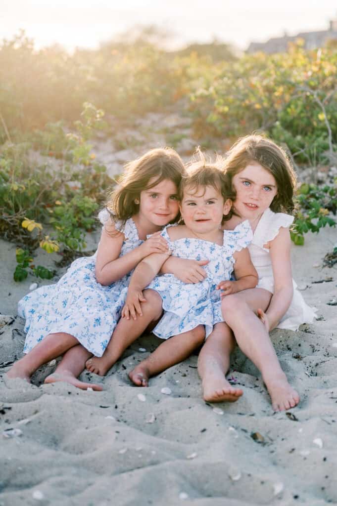 Three young children sit together on a sandy beach, surrounded by greenery. Captured by photographer Kaity Farrell, this Nantucket summer family photo shows two girls in blue floral dresses and one in white, all looking at the camera.