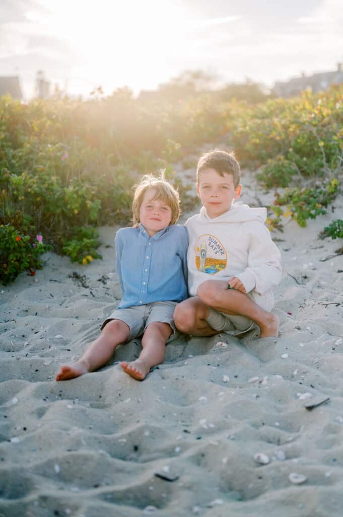 Two young children sit barefoot on sandy ground in front of green bushes, with sunlight shining behind them. Captured by photographer Kaity Farrell, this moment reflects the warmth found in Brant Point Lighthouse family photos.