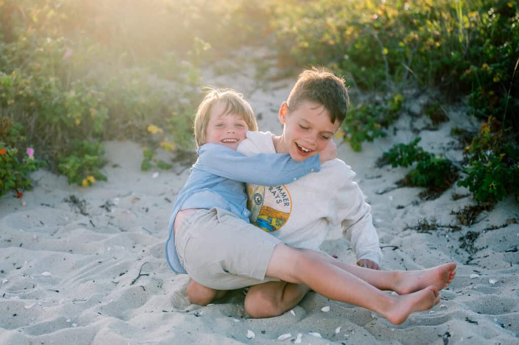 Two young children play and laugh together on sandy ground with greenery behind them—captured by photographer Kaity Farrell, specializing in family portraits near Brant Point Lighthouse.