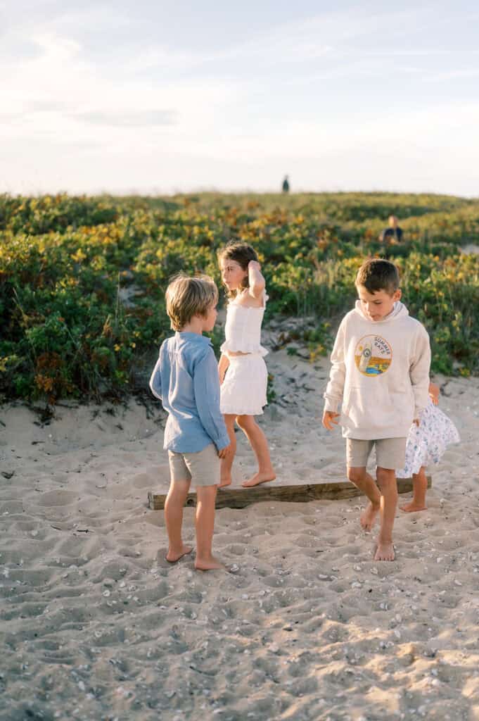 Three children stand barefoot on a sandy beach near a log, with grass and vegetation in the background under a partly cloudy sky—beautiful family portraits near Brant Point Lighthouse by photographer Kaity Farrell.