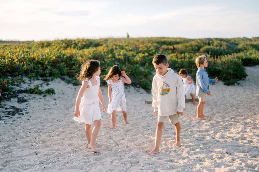 Five children walk and play barefoot on a sandy beach with greenery in the background under a partly cloudy sky during a Nantucket extended family portrait session by photographer Kaity Farrell.