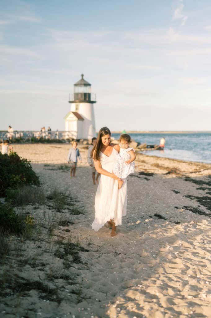 A woman in a white dress carries a young child along a sandy beach at sunset near a Nantucket lighthouse, as photographer Kaity Farrell captures family photos with others in the background.