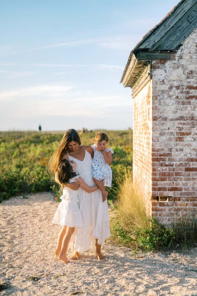 A woman in a white dress stands by a weathered brick building, holding a small child while another holds her hand outdoors on a sunny day during a Nantucket family portrait session.