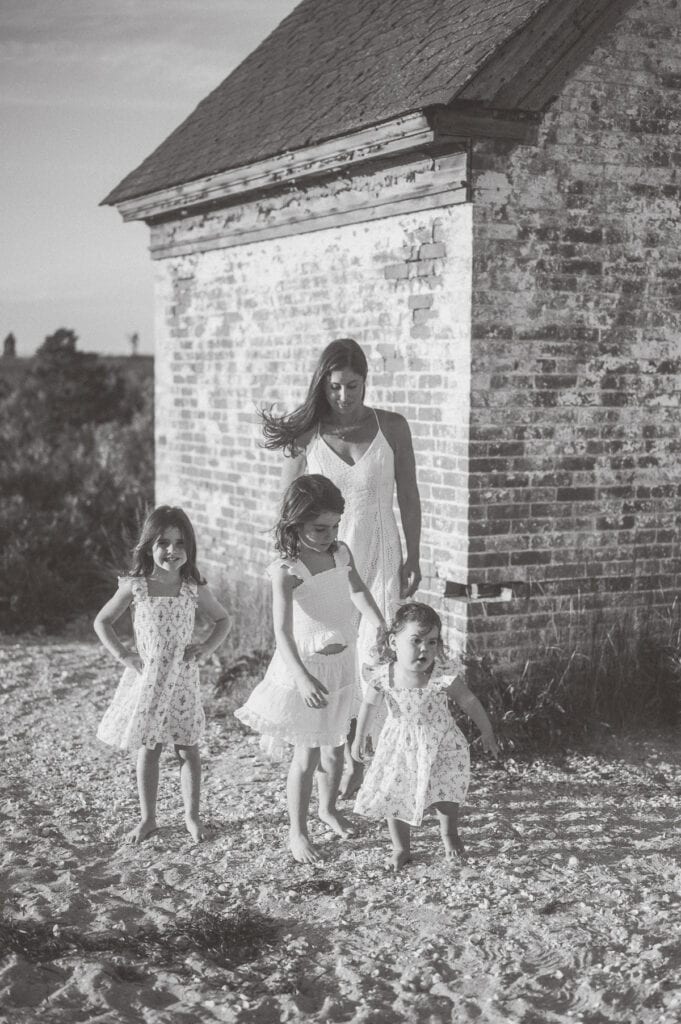A woman and three young girls in white dresses stand outside near a brick building on a sunny day, perfectly capturing Nantucket summer family photos by photographer Kaity Farrell.