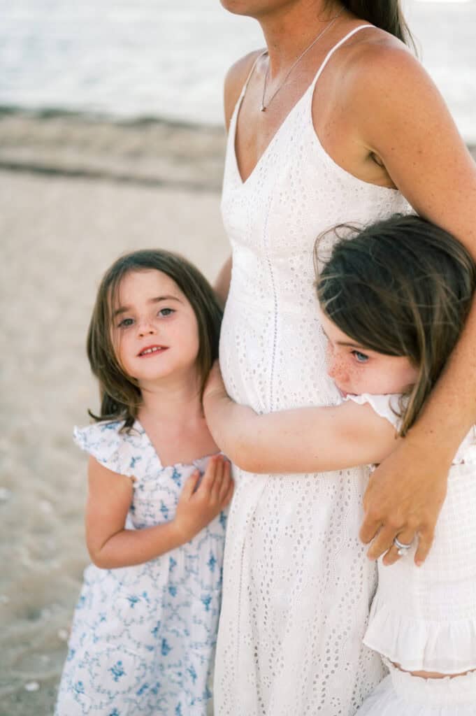 Two young girls stand on a sandy beach hugging a woman in a white dress, with the ocean behind them, beautifully captured by photographer Kaity Farrell during family portraits near Brant Point Lighthouse.
