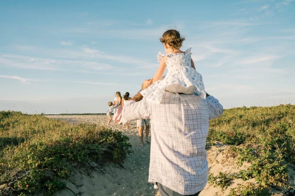 A man carries a young girl on his shoulders while walking along a sandy path; other people, captured by photographer Kaity Farrell, walk ahead under the clear Nantucket sky.