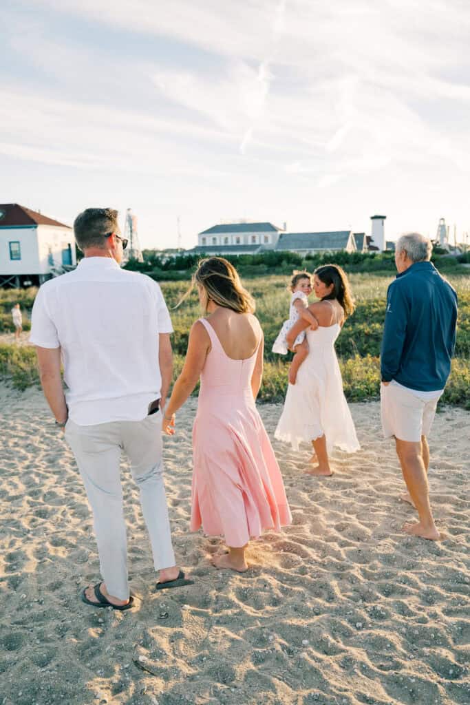 Four adults and a baby walk together on a sandy path near grassy dunes, with houses and a clear sky in the background—perfect for extended family photos Nantucket by photographer Kaity Farrell.