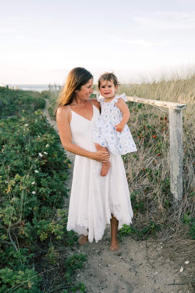 A woman in a white dress stands on a sandy path, holding a crying toddler in a white floral dress. Grasses and bushes border the path under a clear sky—an authentic moment captured by photographer Kaity Farrell for Nantucket family photos.