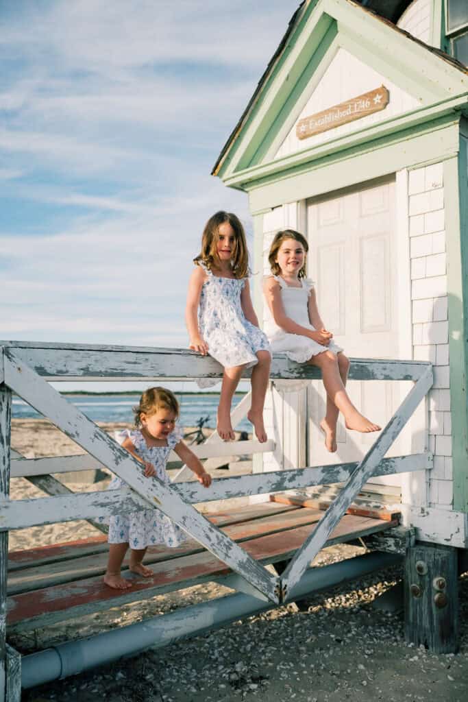 Three young girls in light summer dresses play on a weathered wooden fence next to a small, white beach hut on a sunny day—an idyllic scene captured by photographer Kaity Farrell.