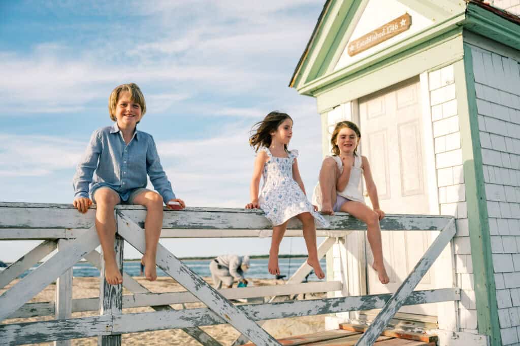 Three children sit barefoot on a white wooden fence at the beach beside a small white building with a sign, under a blue sky, perfectly capturing the essence of Brant Point beach family photos by photographer Kaity Farrell.