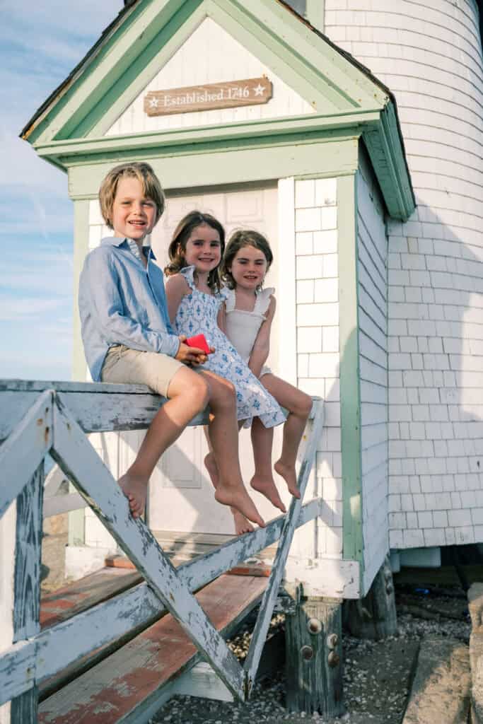 Three children sit barefoot on a wooden railing in front of a white shingled building with a sign reading “Established 1746,” captured by photographer Kaity Farrell for Nantucket vacation family photos.