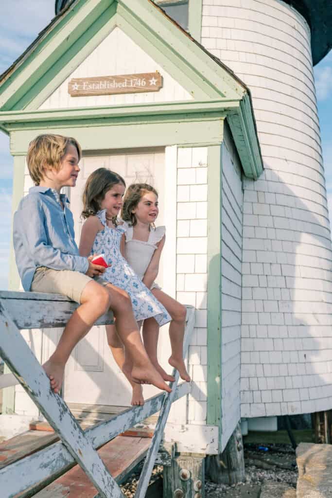 Three children sit barefoot on a white wooden railing in front of Brant Point lighthouse with a sign reading "Established 1746," perfectly capturing the timeless charm of Nantucket family photos by photographer Kaity Farrell.