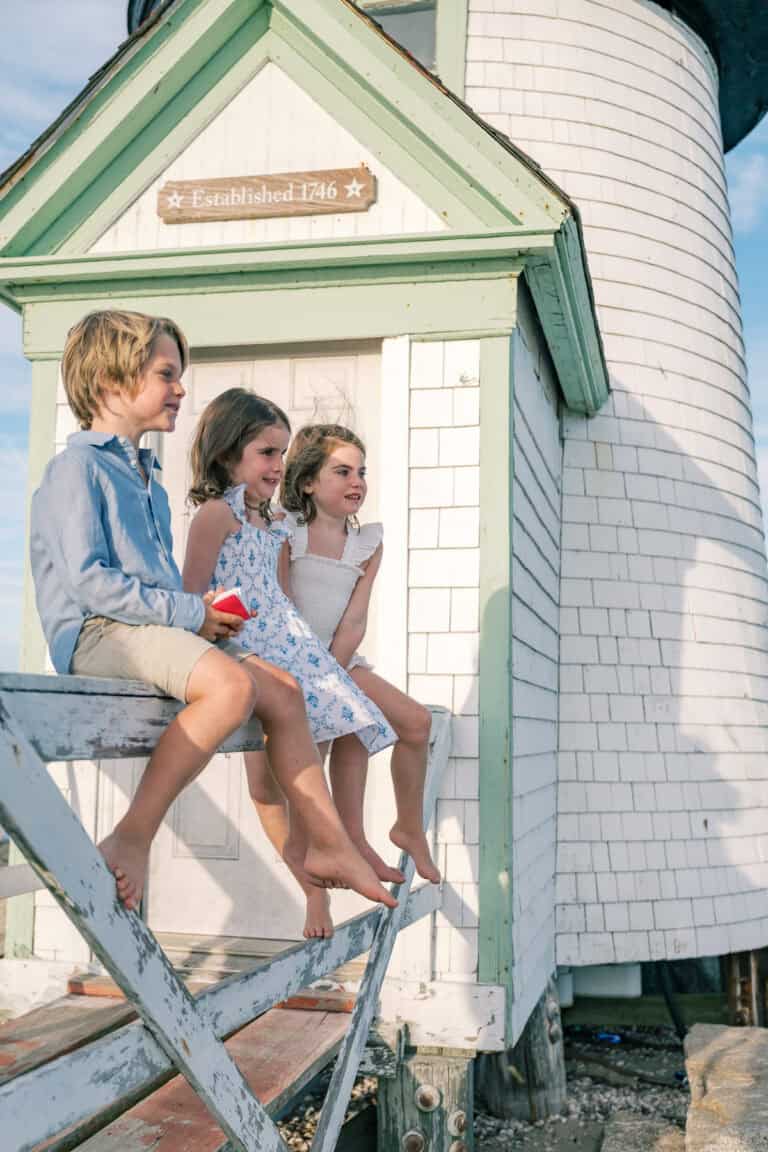 Three children sit barefoot on a white wooden railing in front of Brant Point lighthouse with a sign reading "Established 1746," perfectly capturing the timeless charm of Nantucket family photos by photographer Kaity Farrell.