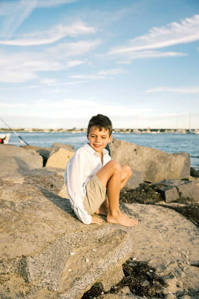 A young boy in a white shirt and khaki shorts sits barefoot on large rocks by the water, with a blue sky and scattered clouds in the background—capturing the essence of Nantucket summer family photos by photographer Kaity Farrell.