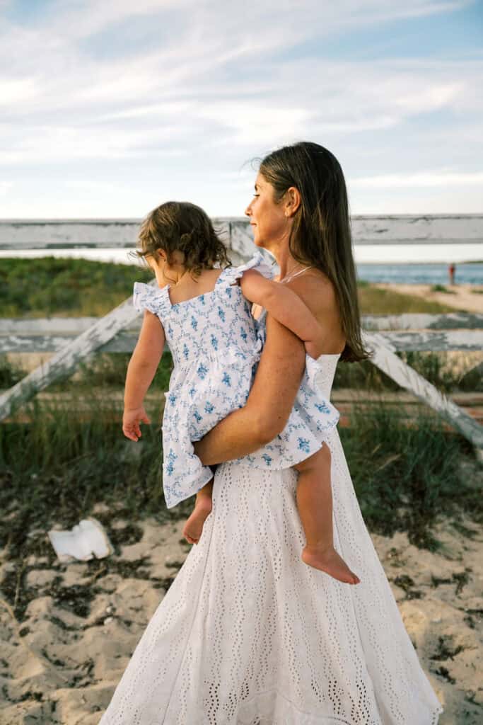 Woman in a white dress holding a young girl in a matching blue and white dress, standing on a sandy path near the beach with greenery and water—captured by photographer Kaity Farrell for Nantucket beach family photos.
