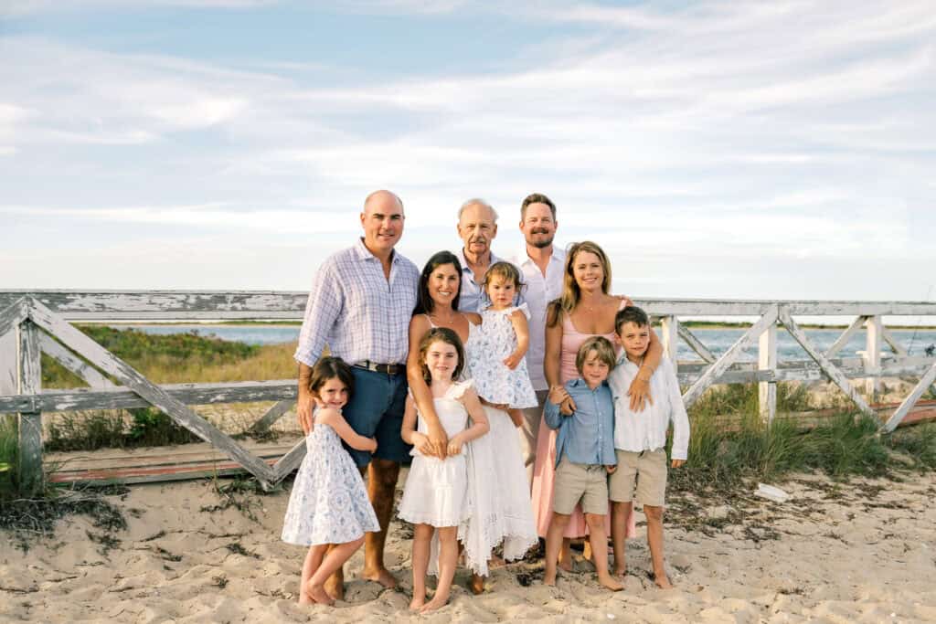 Three adult couples and five children pose together on a sandy beach in front of a wooden fence, with grass and ocean in the background—captured by photographer Kaity Farrell for Brant Point Lighthouse family photos.