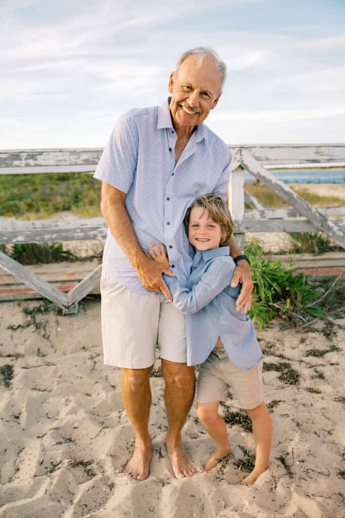 An older man and a young boy stand barefoot on Brant Point beach, smiling at the camera with a wooden fence and grassy dunes in the background—captured by photographer Kaity Farrell.