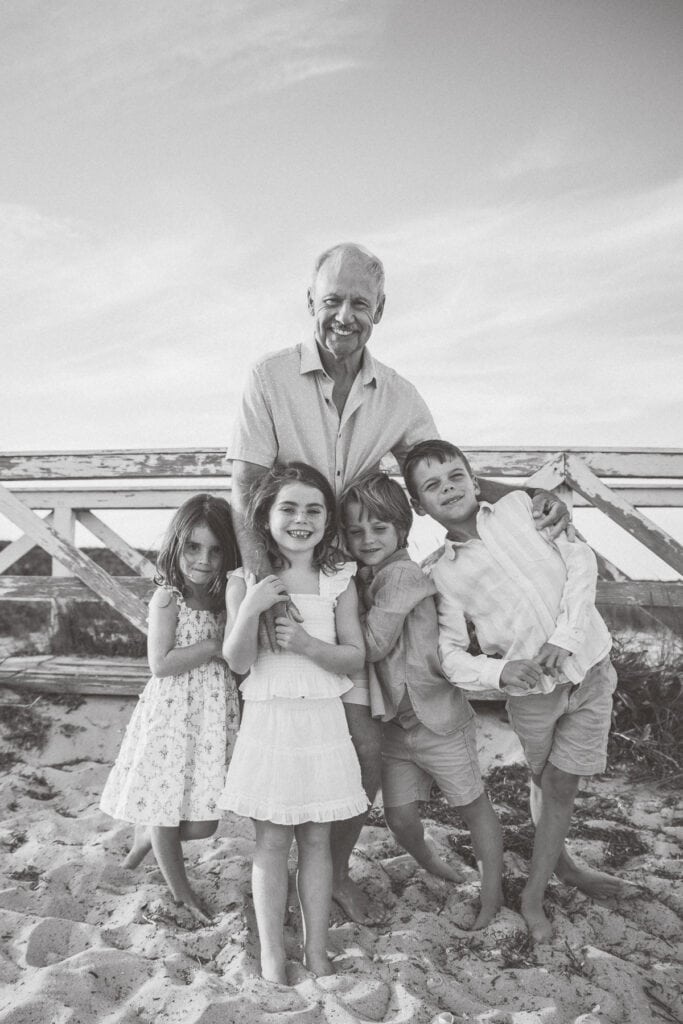 An older man stands on sand with four smiling children in front of a wooden fence at the beach during a Nantucket family portrait session by photographer Kaity Farrell.