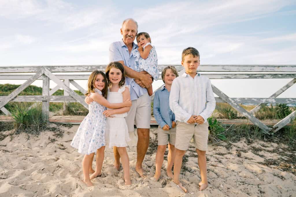 An older man stands on a sandy beach with five children, all barefoot and dressed in light-colored summer clothes—perfect for extended family photos Nantucket style, captured by photographer Kaity Farrell with a wooden fence and blue sky behind them.