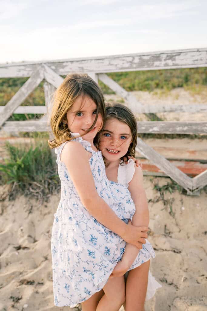 Two young girls in light dresses stand on sandy ground by a weathered wooden fence, with one girl holding the other. Captured by photographer Kaity Farrell, this Nantucket family portrait session features grass and sky in the background.