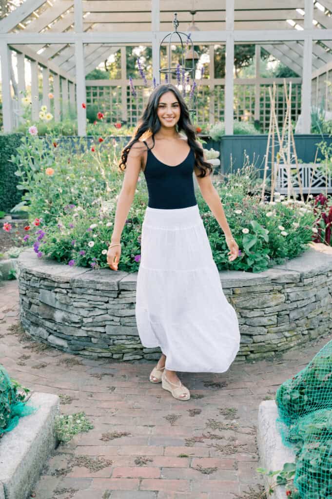 A woman in a black tank top and white skirt stands smiling in a garden, surrounded by flowers and stone planters under a glass greenhouse roof—captured by photographer Kaity Farrell for Nantucket senior portraits.