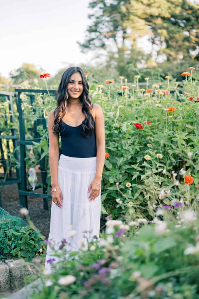 A woman stands in a garden wearing a black tank top and a long white skirt, surrounded by green plants and orange flowers—an elegant scene captured by photographer Kaity Farrell for Nantucket senior portraits.