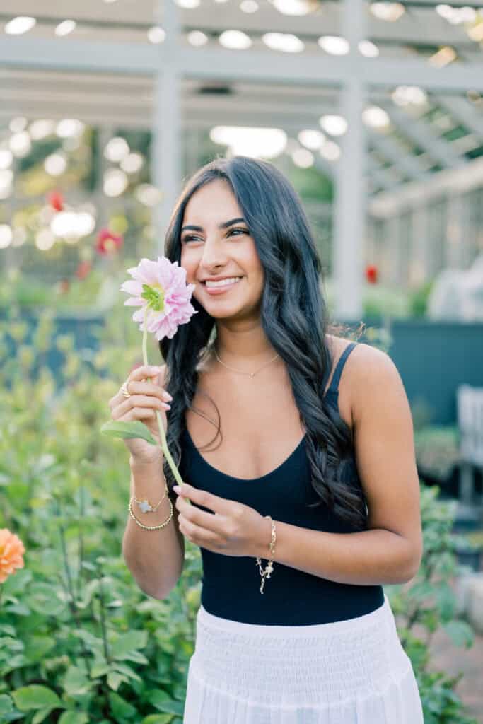 A young woman with long dark hair, wearing a black tank top and white skirt, stands in a garden holding a pink flower and smiling, beautifully captured by Nantucket senior photographer Kaity Farrell.