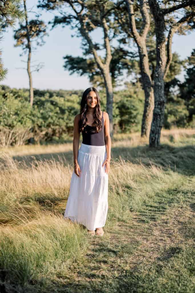 A woman in a black top and long white skirt stands on a grassy path surrounded by trees and sunlight, beautifully captured by photographer Kaity Farrell for Nantucket senior portraits.