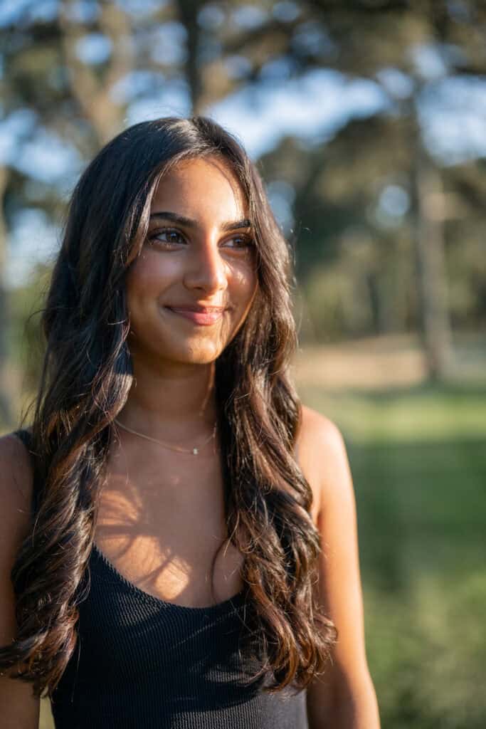 A woman with long, dark hair stands outdoors in sunlight, wearing a sleeveless black top. Captured by photographer Kaity Farrell, this Nantucket senior portrait features blurred trees and greenery in the background.