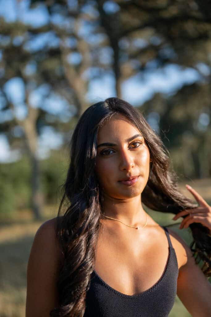 A woman with long dark hair wearing a black sleeveless top stands outdoors in sunlight, trees blurred behind her. This portrait by photographer Kaity Farrell captures the essence of Nantucket senior portraits.