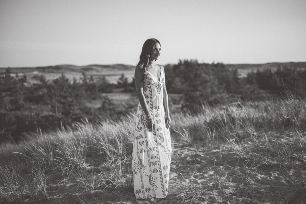 A woman in a long, patterned dress stands in tall grass on sand dunes, with trees and distant hills in the background. Captured by photographer Kaity Farrell, this black and white image channels the timeless feel of Surfside Beach senior photos.