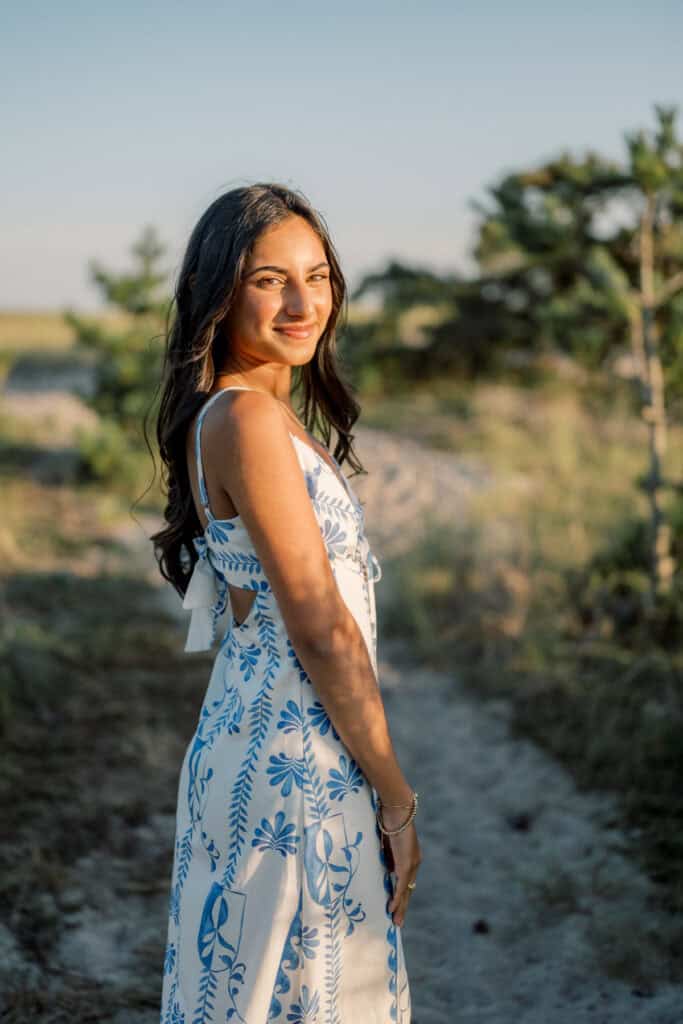 A woman in a white sundress with blue floral patterns stands on a sandy path outdoors, smiling at the camera with greenery in the background—captured by photographer Kaity Farrell for Nantucket beach senior portraits.