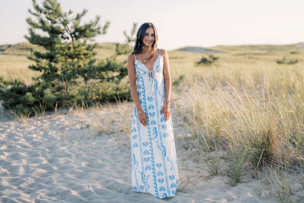 Woman in a long, white and blue floral dress stands on sandy ground with tall grass and pine trees in the background, captured by photographer Kaity Farrell during a Surfside Beach senior photos session.