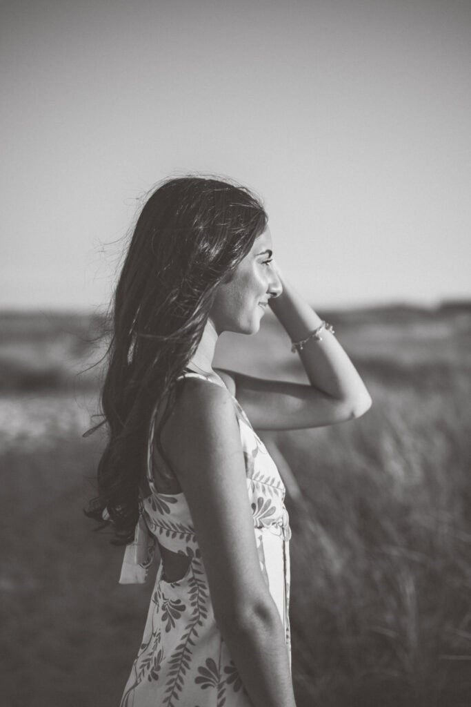 A woman with long hair stands outdoors in a patterned dress, looking to the side and touching her hair; the scene is in black and white, captured by photographer Kaity Farrell for Nantucket senior portraits.