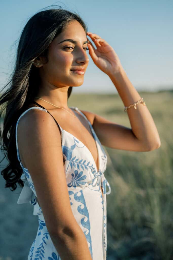 A woman in a sleeveless, patterned dress stands outdoors in sunlight, one hand raised to her forehead, as grassy landscape glows behind her—a signature moment from photographer Kaity Farrell’s Nantucket summer senior session.