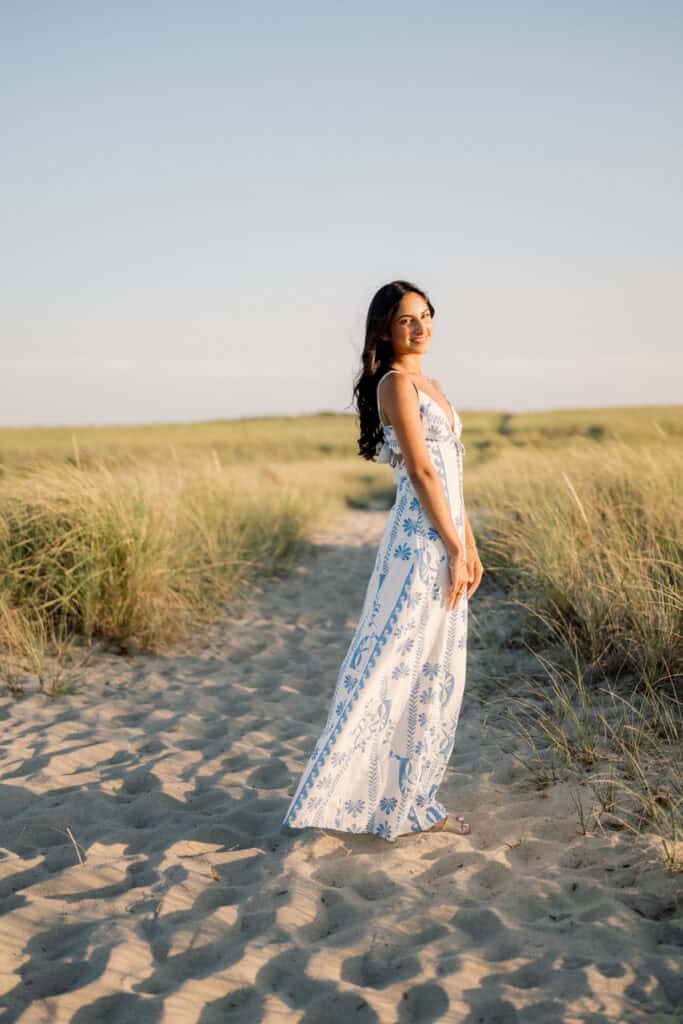 A young woman in a long white and blue patterned dress stands on a sandy path surrounded by tall grass under a clear sky, perfect for Nantucket beach senior portraits photographed by Kaity Farrell.