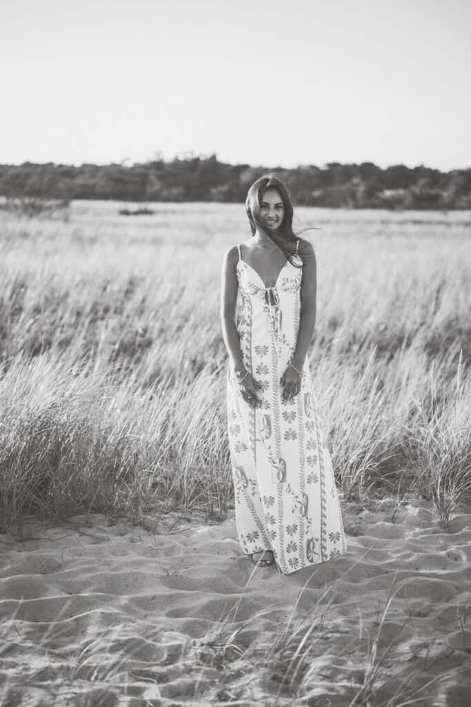 A woman in a patterned maxi dress stands on sandy ground in a grassy field, with trees in the background. Captured in black and white by photographer Kaity Farrell, this image evokes the timeless elegance of Nantucket senior portraits.
