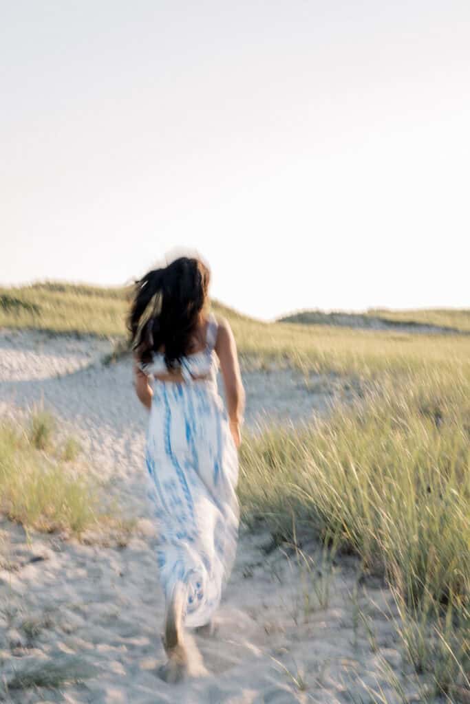 A woman in a flowing white and blue dress walks barefoot along a sandy path through grassy dunes under a bright sky, capturing the essence of Nantucket beach senior portraits by photographer Kaity Farrell.