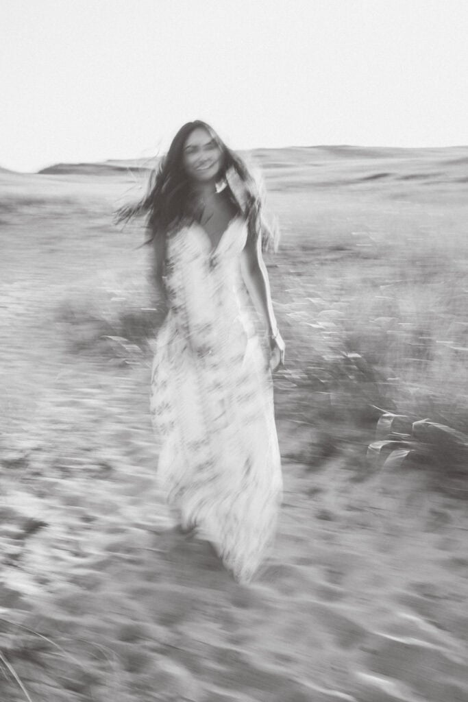 A blurred black and white photo by photographer Kaity Farrell shows a woman in a long dress walking outdoors on a sandy path with grass around her, capturing the timeless feel of Nantucket senior portraits.