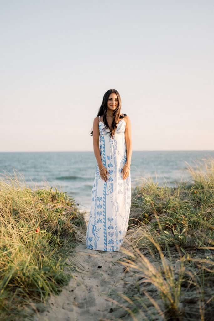 A woman in a long, white and blue floral dress stands on a sandy path amid grass by the ocean under a clear sky, captured during a Nantucket summer senior session by photographer Kaity Farrell.
