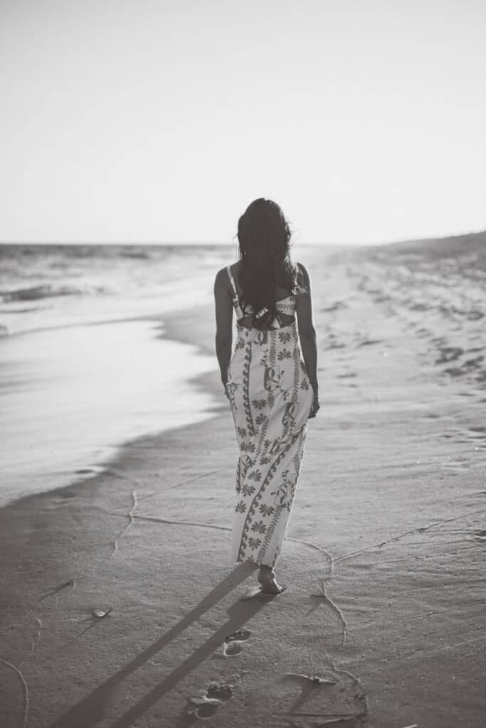 A woman in a patterned dress walks barefoot along a sandy beach, leaving footprints behind her. Captured in black and white by photographer Kaity Farrell, this could be the perfect inspiration for Surfside Beach senior photos.