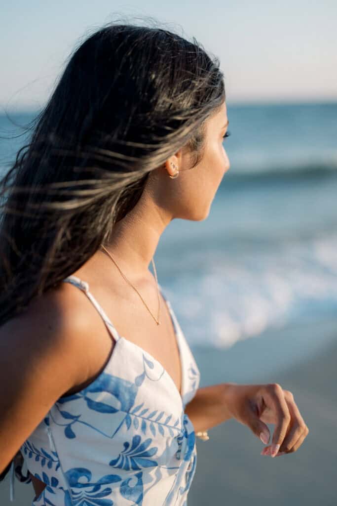 Woman with long dark hair wearing a blue and white dress stands on a beach, looking toward the ocean, with waves in the background—a serene Nantucket senior portraits moment by photographer Kaity Farrell.