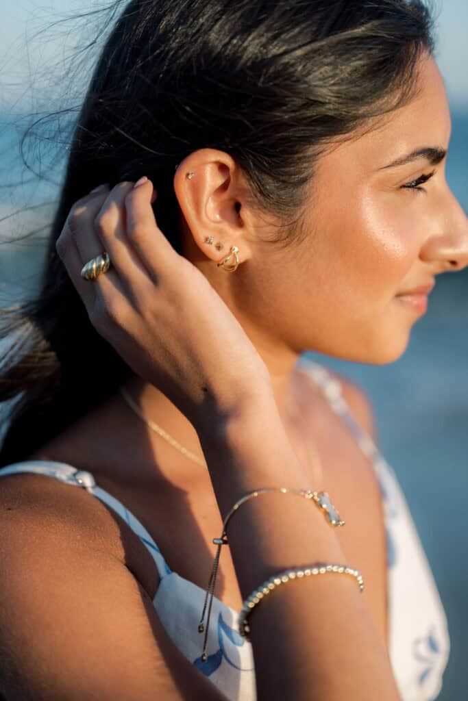 Woman with smooth skin and long dark hair wears gold jewelry, including earrings, rings, bracelets, and a necklace, while touching her ear and looking to the right in natural light—captured by photographer Kaity Farrell.