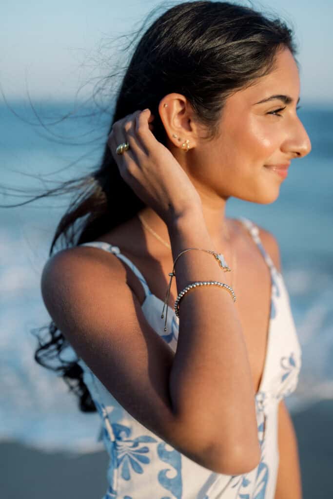 A woman with long dark hair, wearing a patterned dress and gold jewelry, stands by the ocean with her hand near her ear.