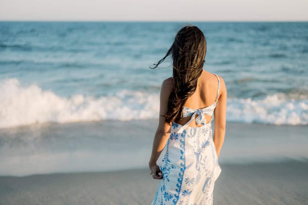 A woman with long dark hair in a white and blue dress stands on the beach facing the ocean, with gentle waves in the background, captured by Nantucket senior photographer Kaity Farrell.